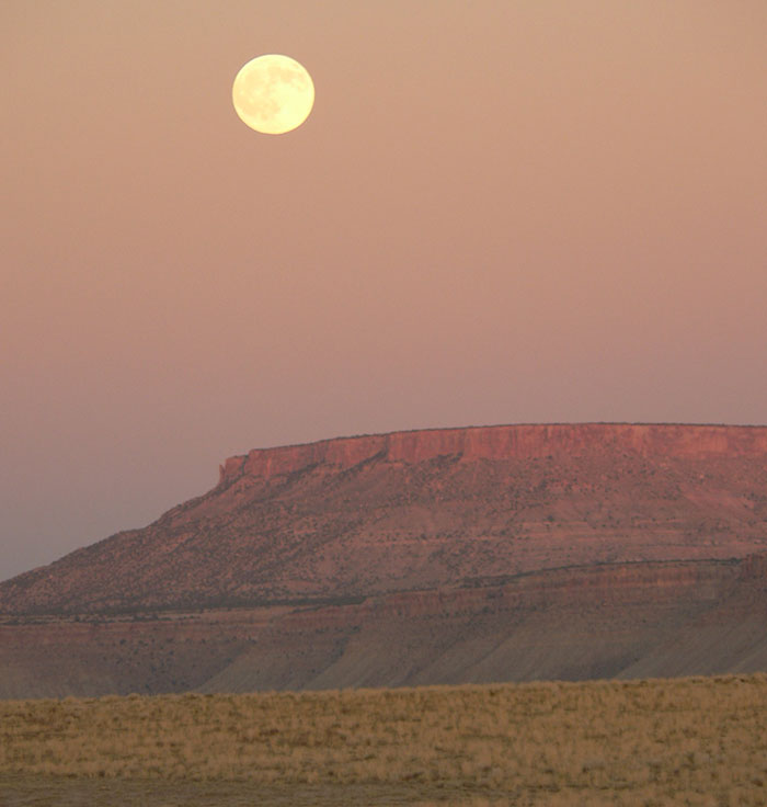A full moon hanging above a vast, reddish desert mesa, illustrating a landscape from the US States.