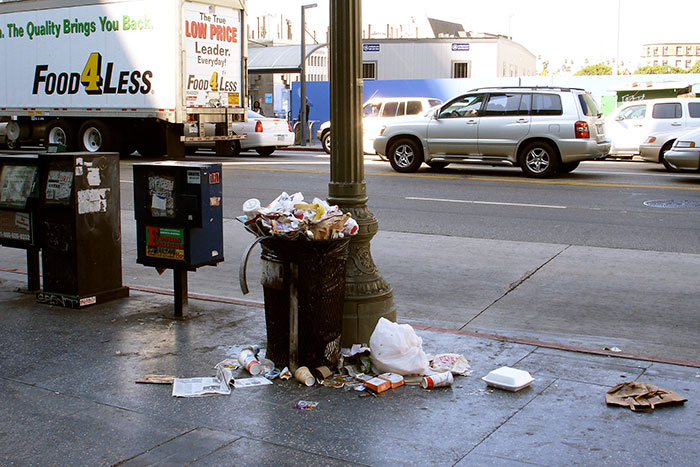 A street scene with overflowing trash cans and passing cars, showing less glamorous US states.