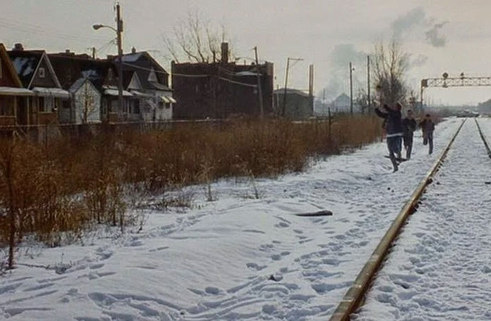 Three figures running along snow-covered train tracks through an industrial area, a scene from US states glorified in movies.