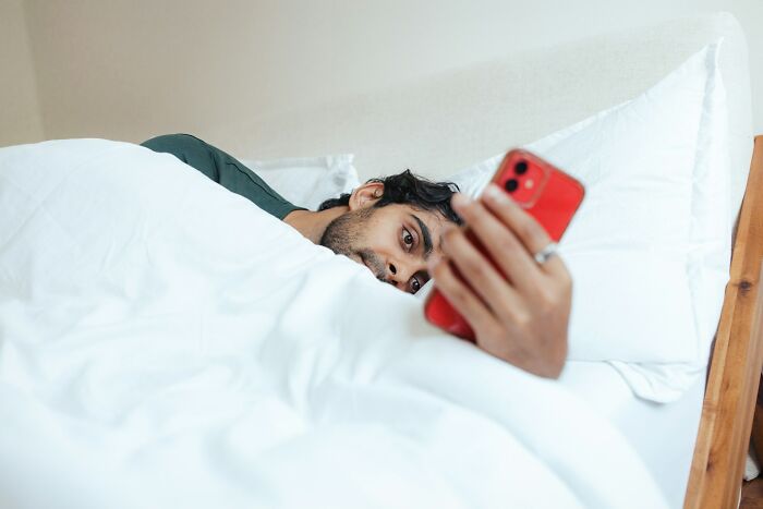 A man in bed, covered in white sheets, looking at a red smartphone. His expression suggests contemplation or concern. He's one of the men suffering.