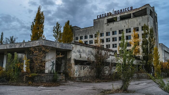 An abandoned hotel in Pripyat with faded Russian lettering on top, surrounded by overgrown trees under a grey sky. Disturbing d***h stories.
