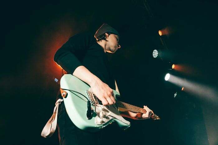 A musician on stage, lit by spotlights, playing a light green electric guitar. Celebrity relatives face unique costs.