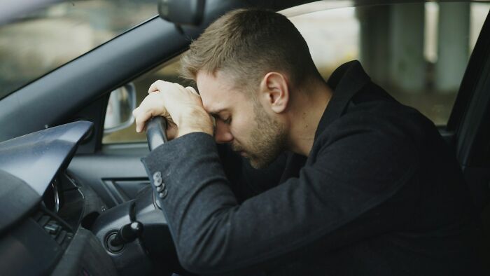 A man with short brown hair and a beard, in a black coat, resting his head on his hands on a car steering wheel, looking distraught. Engagement stories can have moments of chaos.