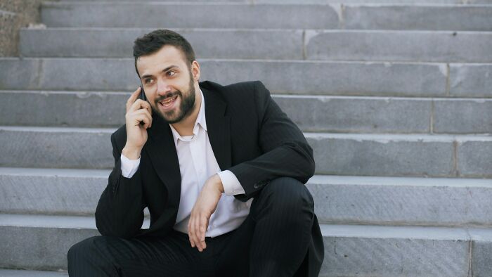 A smiling, bearded man in a suit talking on a cell phone, sitting on concrete stairs, embodying silent luxuries.
