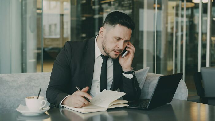 A lawyer in a black suit, on the phone, taking notes in a notebook, with a laptop and coffee. Hardest cases.