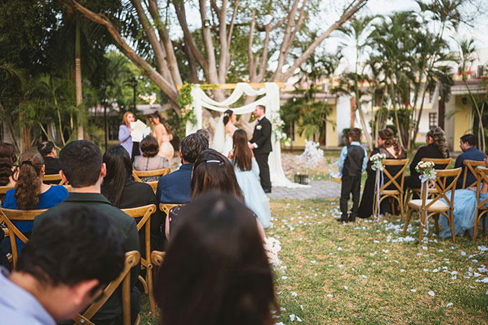Outdoor wedding ceremony with guests seated, witnessing a marriage under an archway, surrounded by greenery.