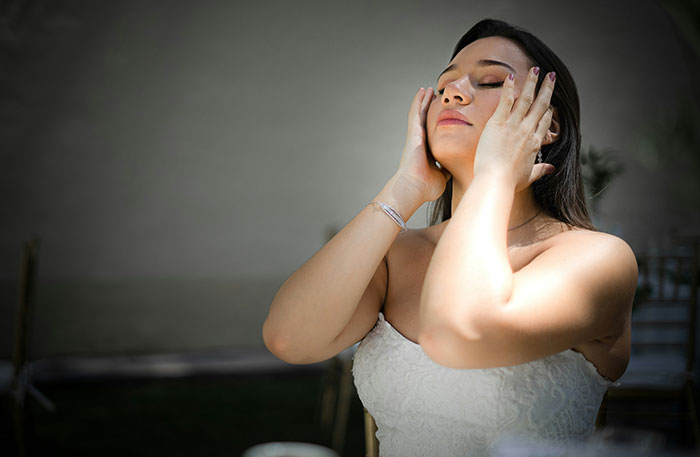A bride with closed eyes, hands on face, experiencing a wedding moment. Sunlight illuminates her, creating a stressed or relieved look.