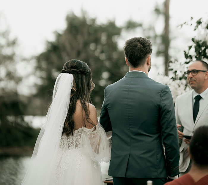 A bride and groom facing a wedding officiant outdoors. The main focus is on the back of the wedding couple.