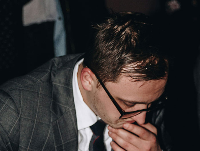 Man in glasses and suit, hand covering mouth, likely reacting to a funny or surprising wedding moment.