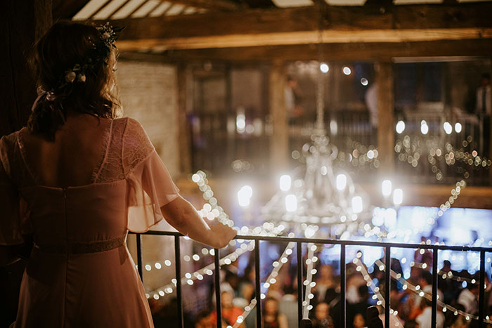 A woman in a pink dress and floral headpiece views a lively wedding celebration below. Wedding moments.