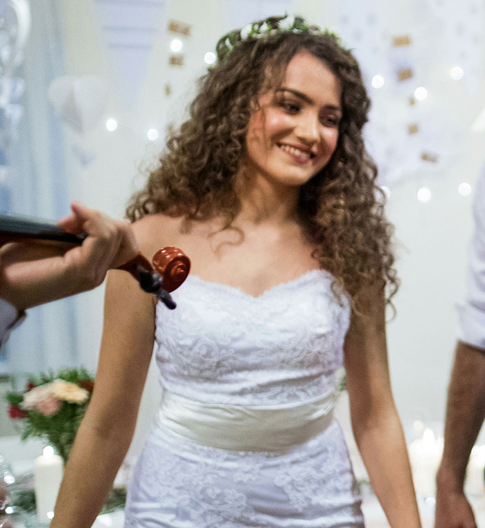 A young woman with curly hair, wearing a white strapless wedding dress and flower crown, smiling while a violin plays. Wedding moments.