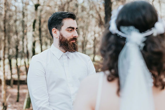 A bearded groom in a white shirt looking at his bride during their wedding in a forest. This marriage moment captures the expectation.