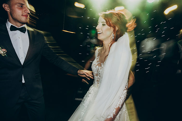 A happy wedding couple dancing, with the groom in a suit and the bride in a white dress, at a lively reception.