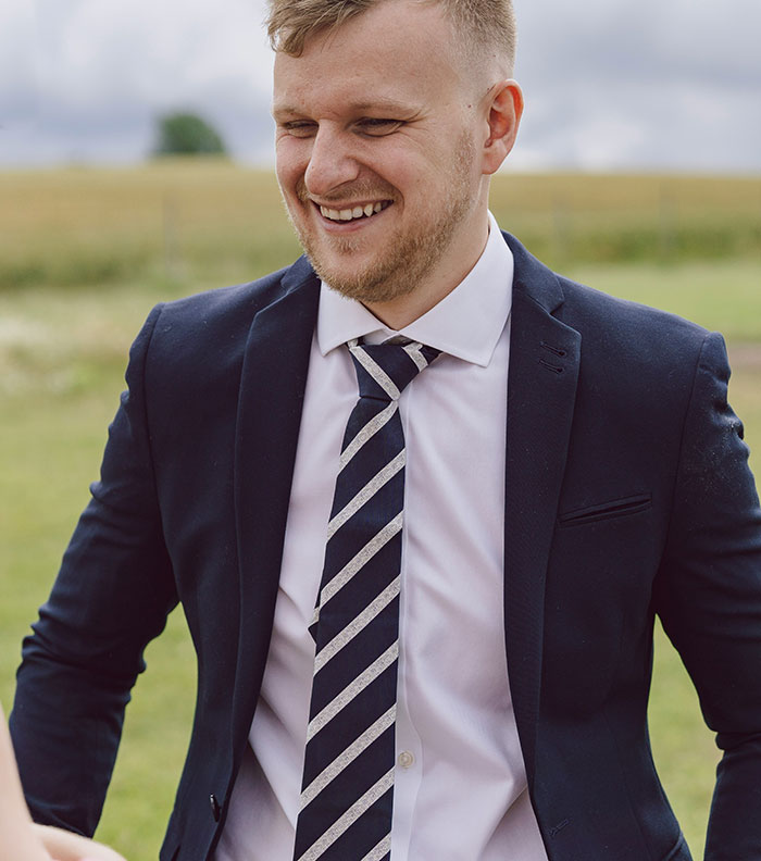 A smiling man in a suit and striped tie, enjoying a wedding moment outdoors.