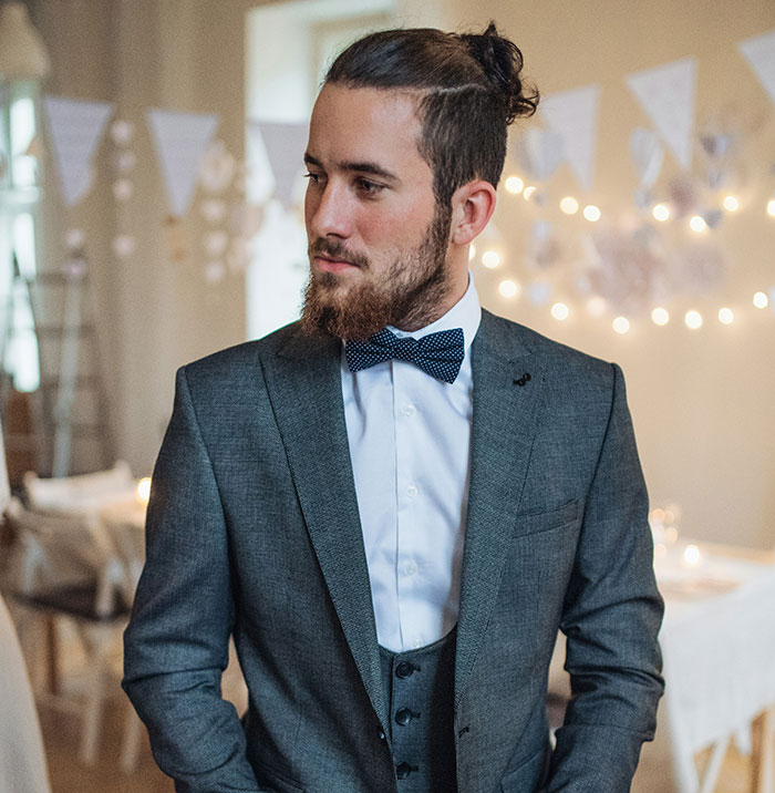 A man with a beard and man bun, dressed in a grey suit and polka-dot bow tie, at a wedding.