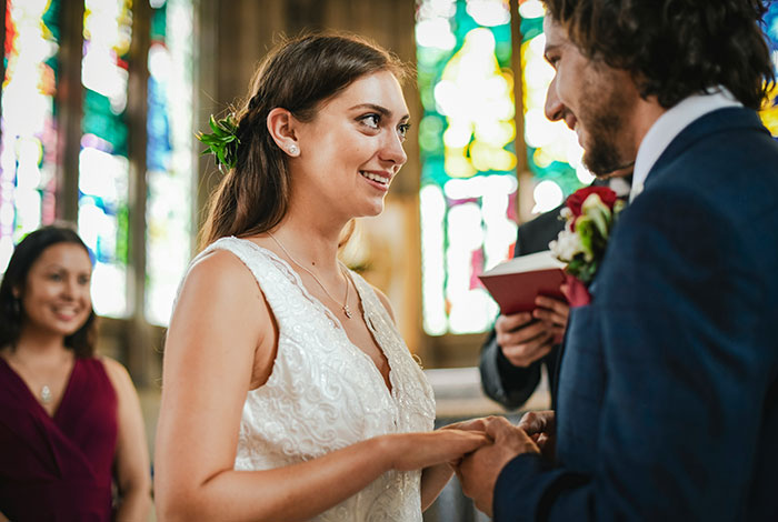 A smiling bride and groom exchanging vows, holding hands during their wedding ceremony, with stained glass windows in the background.