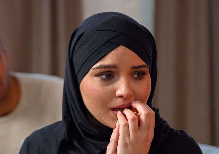 A wife becomes Muslim, wearing a black hijab, anxiously biting her nails, looking concerned.