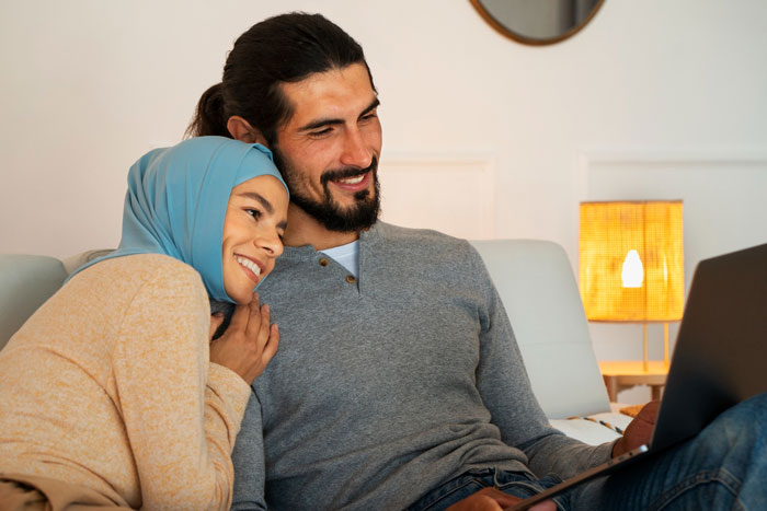 A wife becomes Muslim and smiles beside her husband, who is holding a laptop. The couple sits closely, embracing.