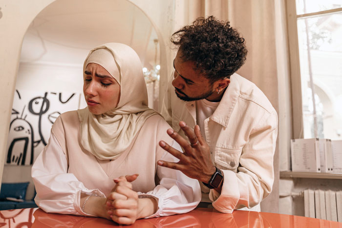 A distressed wife, wearing a hijab, sits with clasped hands as her husband talks to her. Wife becomes Muslim.