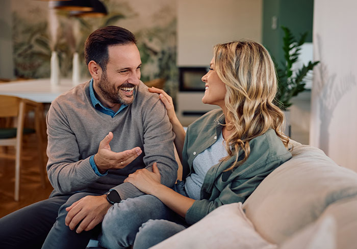 A smiling man and woman sit on a sofa, the woman's hand on the man's shoulder. He talks to his wife about her job.