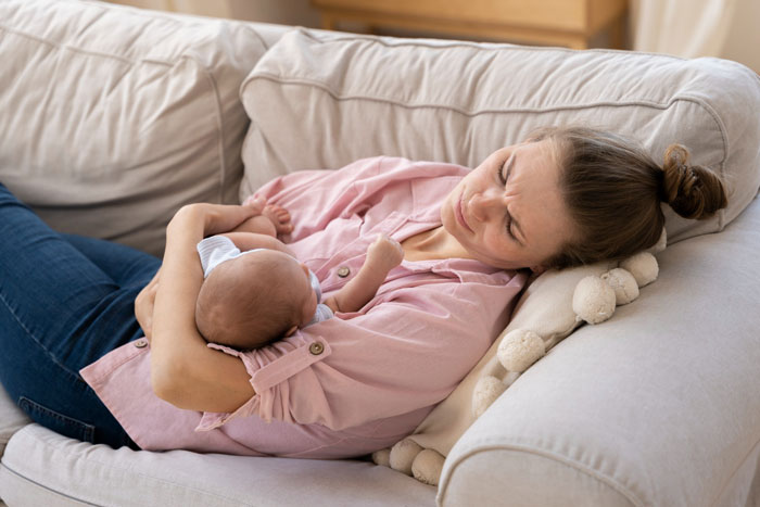 A new mom, a postpartum wife, holding her newborn on a sofa, looking stressed. Her eyes are closed and brow furrowed.