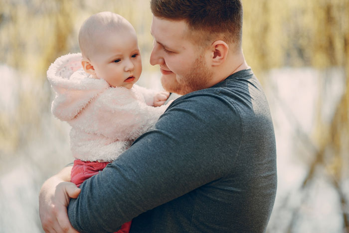 A man in a gray shirt holding a baby in a pink fluffy jacket. The baby looks toward the man, a symbol of postpartum challenges.