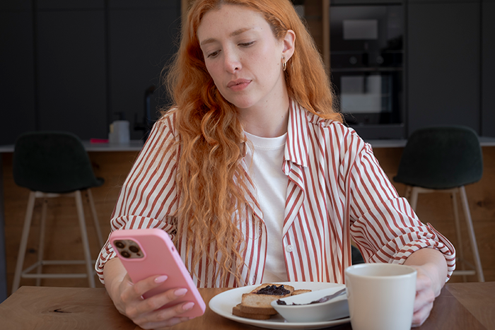 Woman looks at phone sitting by plate with ruined pasta and cup in kitchen