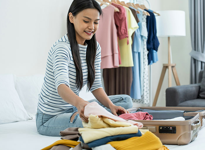 Young woman packing a suitcase, smiling happily, with clothes on a rack in the background. Her dream trip is near.