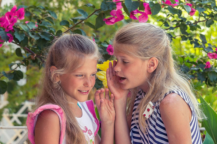 Two young girls whispering in a garden with flowers representing sisterly bond Two young girls whispering in a garden with flowers representing sisterly bond