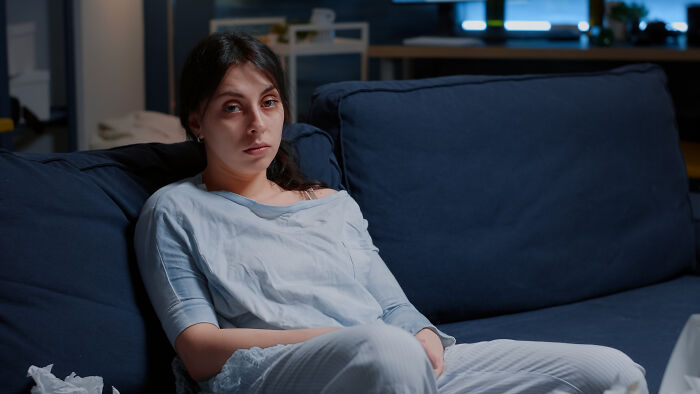 A tired young woman in blue pajamas sits on a navy couch with tissues, highlighting the need for routine cancer screening.