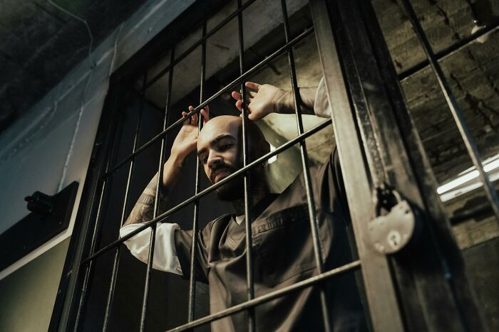 A man with a beard and tattoos, wearing a dark shirt, holding prison bars, looking forlorn and experiencing heartbreak.
