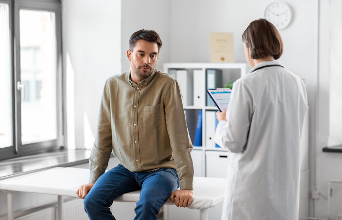 A heartbroken man in a doctor's office, sitting on an examination table while a doctor holds a clipboard, discussing breakups.