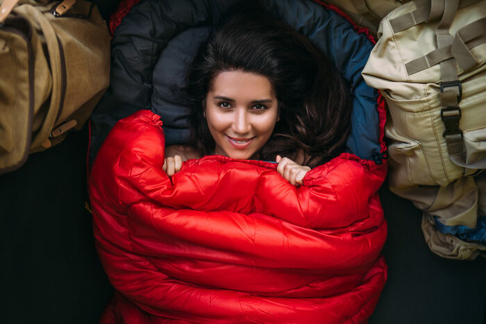 A smiling woman tucked into a red sleeping bag, surrounded by camping gear. She doesn't feel heartbroken about breakups.