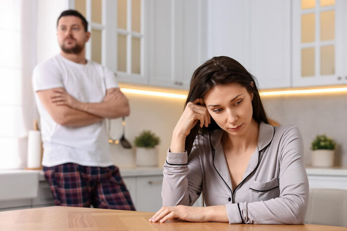 A heartbroken woman sits at a table, looking down, while a man stands in the background, arms crossed, after a breakup.