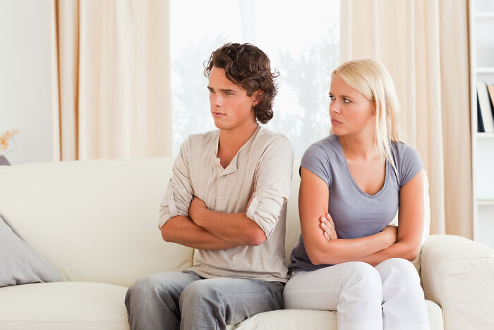 A man and woman sitting on a couch, backs turned, arms crossed, looking angry after a heartbreaking breakup.