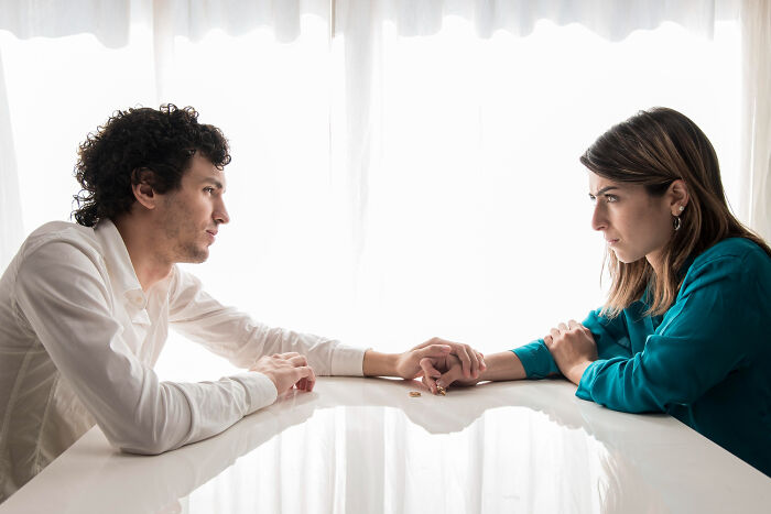 A man and woman looking away from each other, holding hands over wedding rings on a table, experiencing a messy breakup.