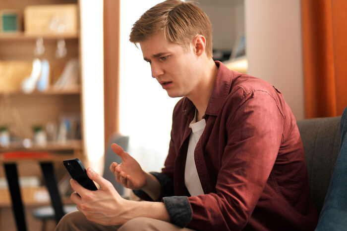 A distraught man sits on a couch, staring at his phone with a heartbroken expression. He is experiencing a breakup.