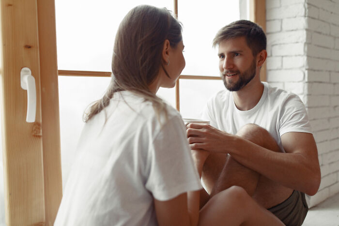 A couple in white shirts sits by a window, talking and holding hands, perhaps discussing their past breakups.