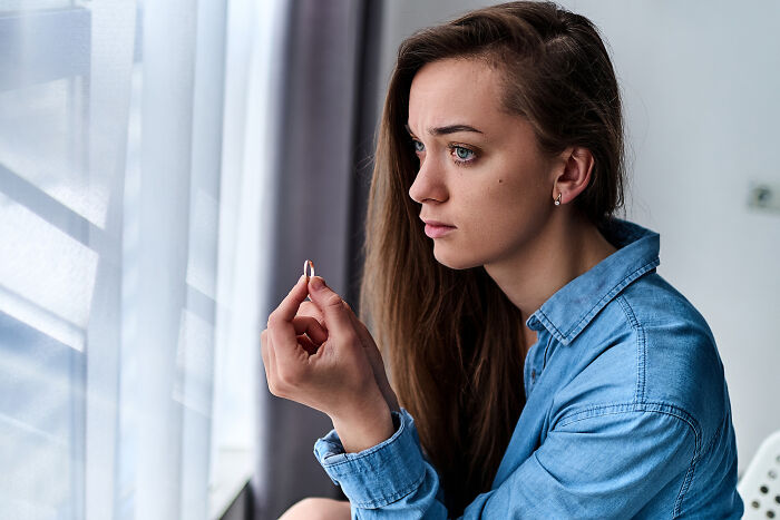 A heartbroken woman in a blue shirt holds a ring, contemplating her unforgettable breakup near a window.