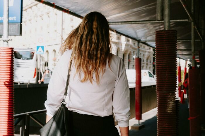 A woman with wavy hair, back to camera, walks under scaffolding in a city, experiencing heartbroken emotions.