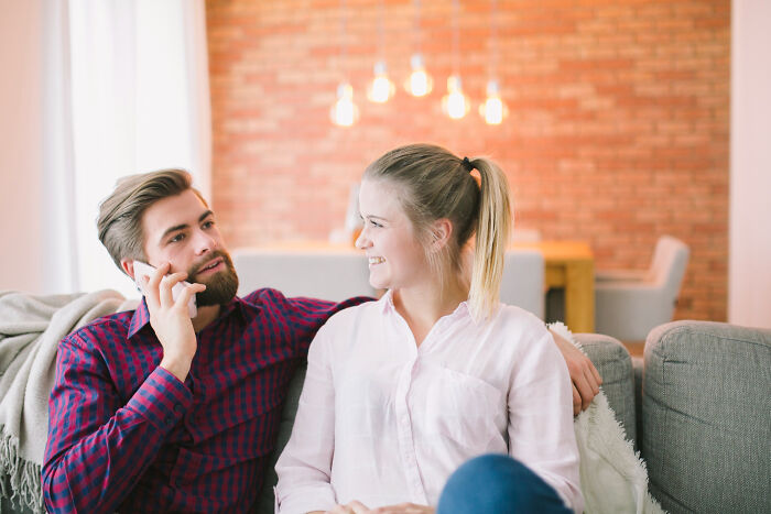 A bearded man on the phone next to a smiling woman on a couch. Could this be about sad breakups?