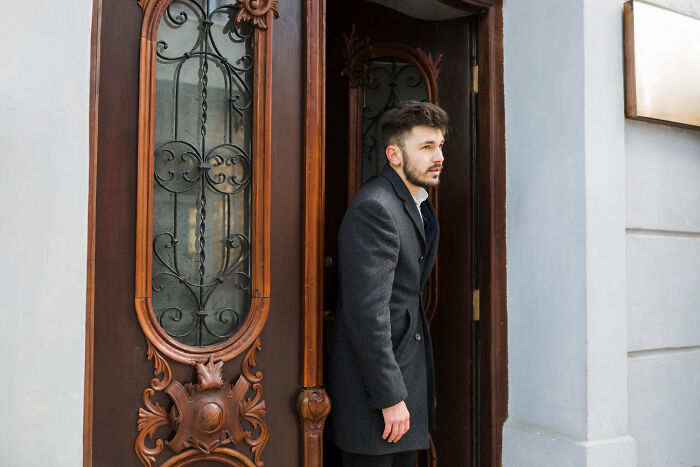 A bearded man in a dark grey coat stands in an ornate doorway, looking away, reflecting on heartbreaking breakups.