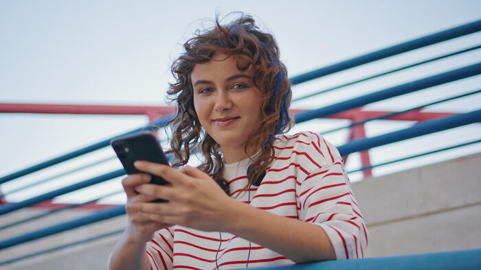 A young woman with curly hair, wearing a striped shirt and headphones, looks at the camera while holding her phone. Breakups.
