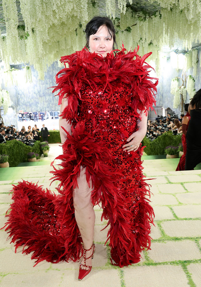 A woman in a red sequined, feathered gown at Met Gala 2026. This worst dressed star wears red studded heels.