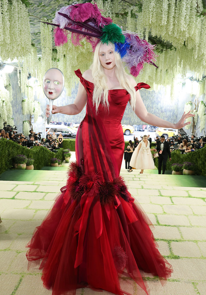 A woman in a red feathered dress and elaborate headpiece, holding a mirror-mask, at the Met Gala 2026. Worst dressed stars.