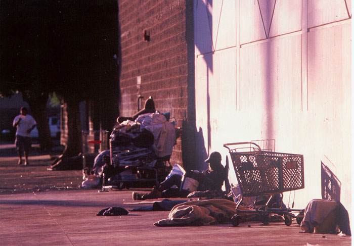 Sidewalk scene showing homeless people and shopping cart near building wall
