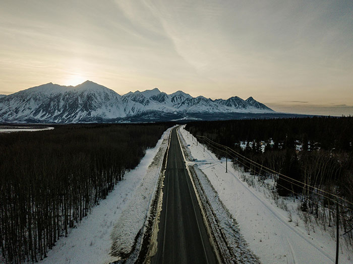 An aerial view of a long, snowy road leading towards mountains at sunset. Evokes drivers' experiences and road rage.