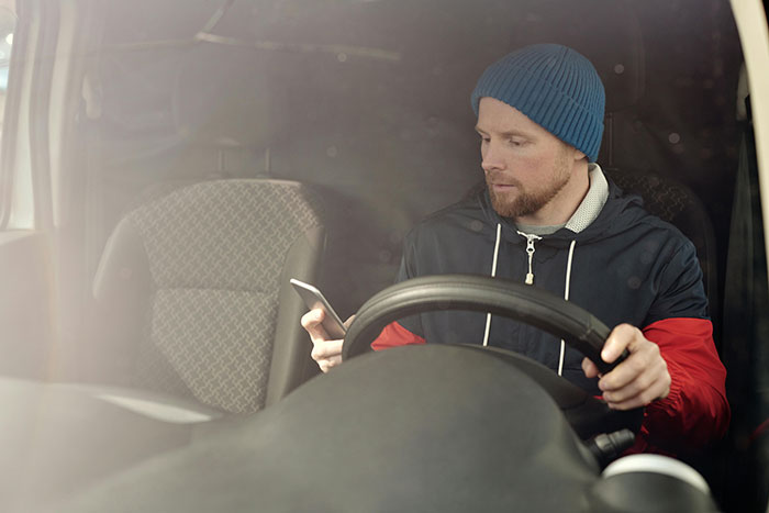 A man in a blue beanie and jacket texting on his phone while driving, exemplifying distracted drivers and road rage.