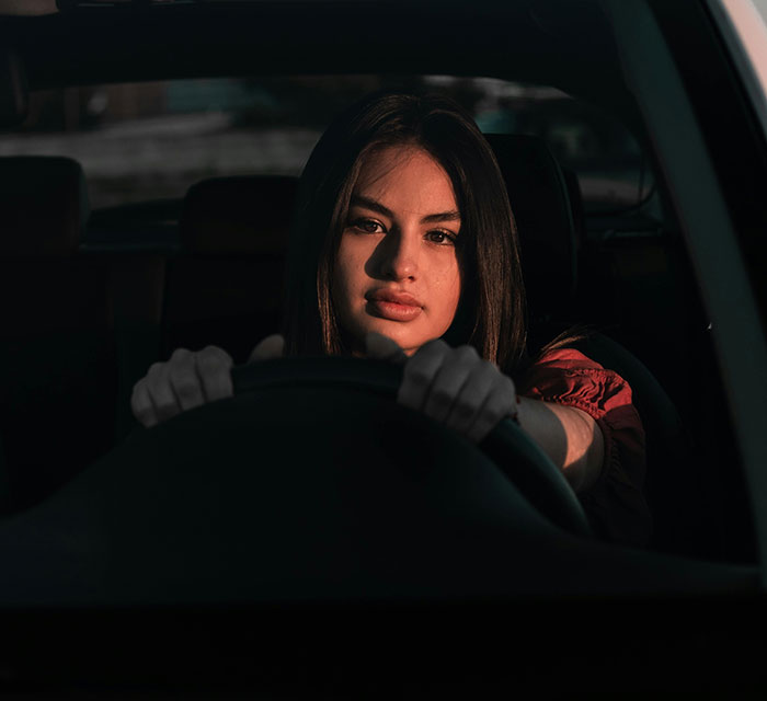 A young woman with dark hair behind the steering wheel, sunlit on her face, depicting the intensity of road rage.