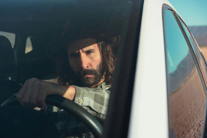 A serious male driver with a beard and hat, looking intensely from behind the wheel. A possible road rage incident.